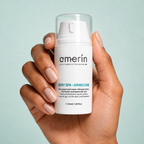Une main aux ongles manucurés tient un flacon-pompe blanc de crème pour les mains anti-âge Amerin sur fond bleu clair.