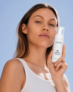 Une femme en débardeur blanc tient un flacon blanc d’amerin ESSENTIAL | Mousse nettoyante enzymatique sur fond de ciel bleu dégagé.