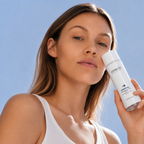 Une femme au teint net tient un flacon blanc AMERIN ESSENTIAL | Eau micellaire à la B5 et à l'aloe vera devant un ciel bleu lumineux.