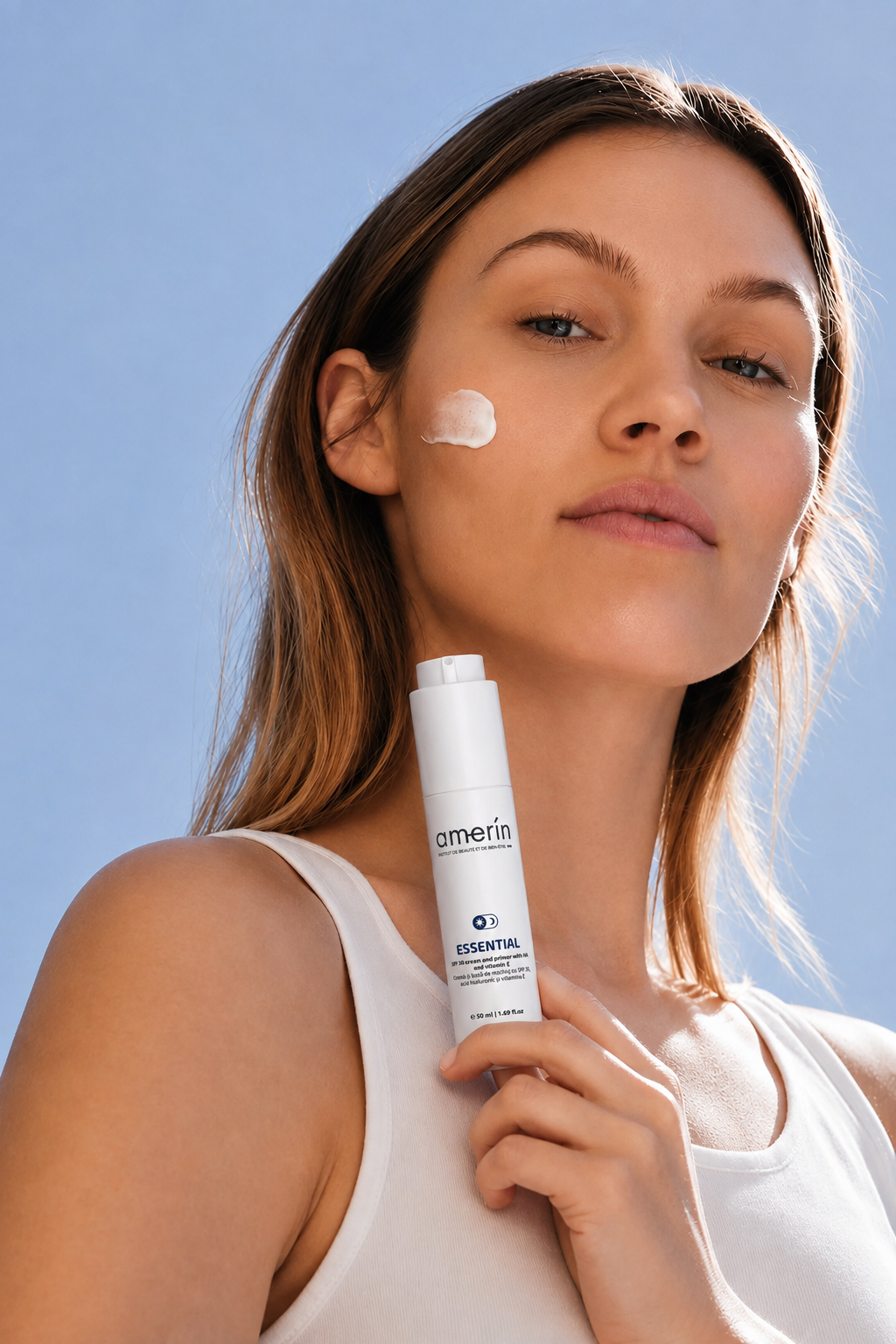 Una donna con una noce di crema bianca sulla guancia tiene un flacone bianco ESSENTIAL | Crema SPF30 e Primer con HA e Vitamina E su sfondo di cielo azzurro sereno.