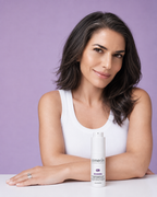 Femme aux cheveux foncés et au débardeur blanc souriant à côté du flacon de 30 ml de sérum raffermissant et repulpant amerin REBORN sur une table blanche, sur fond lavande